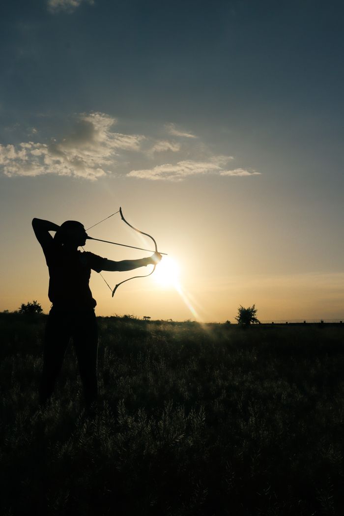 A dramatic silhouette of an archer aiming a bow against a sunset sky.
